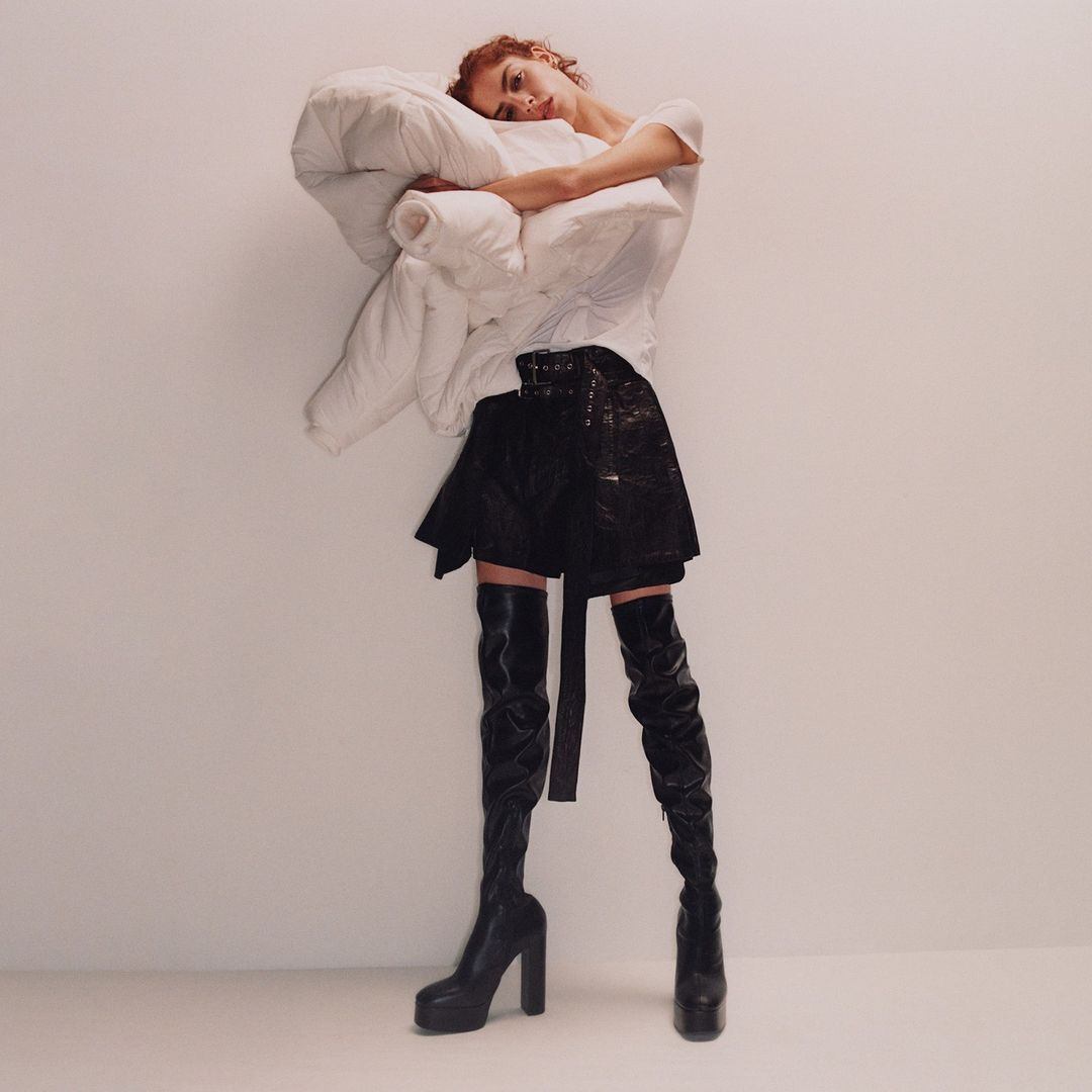 Fashionable woman poses with a large pillow, wearing a white shirt, black mini skirt, and black thigh-high platform boots in studio.