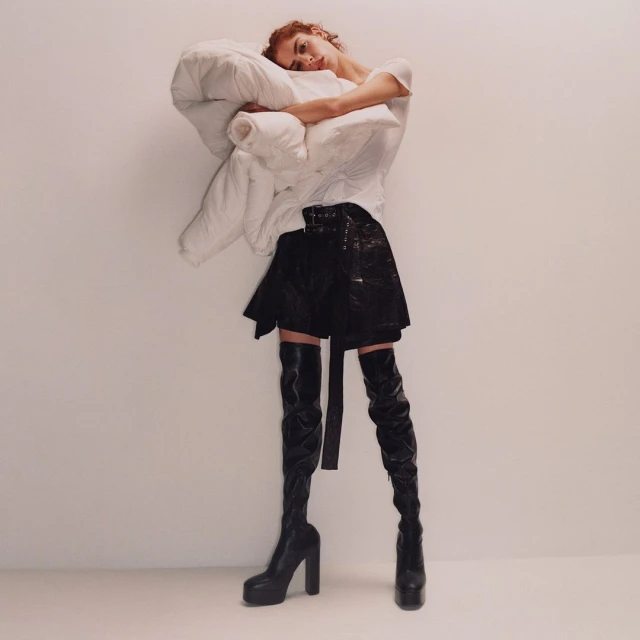 Fashionable woman poses with a large pillow, wearing a white shirt, black mini skirt, and black thigh-high platform boots in studio.