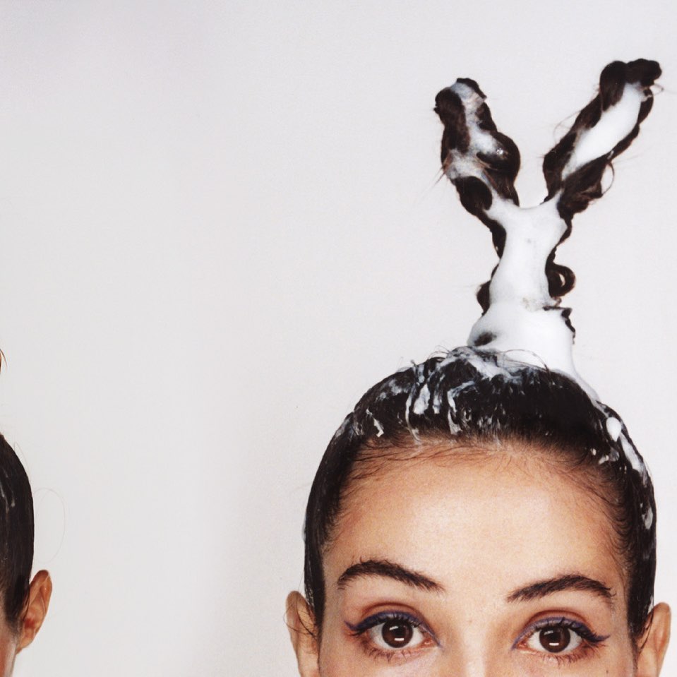 Close-up studio shot of woman's face with soap foam styled in hair; another woman's face is partially visible on the left.