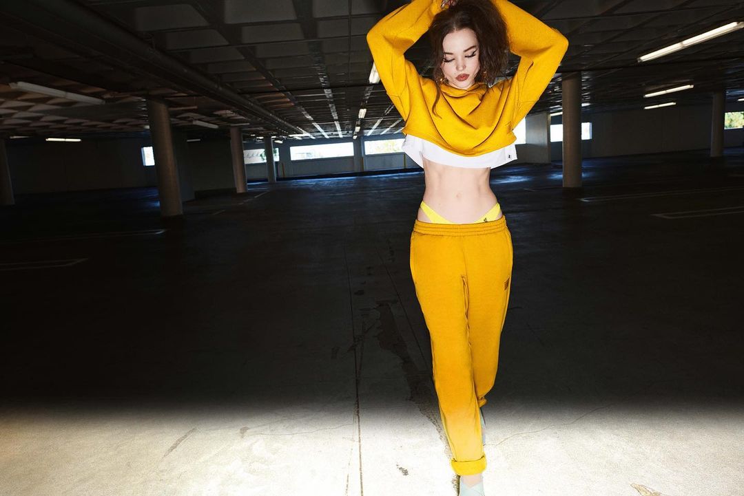 Young woman in yellow sports outfit strikes a playful pose in underground parking garage with diffused lighting and open concrete background.