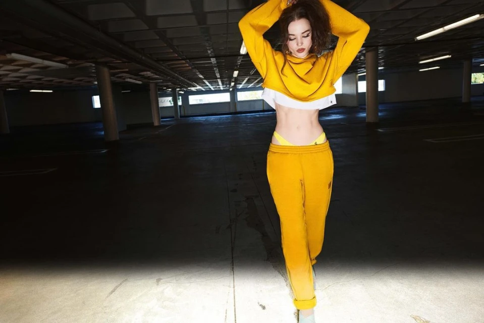 Young woman in yellow sports outfit strikes a playful pose in underground parking garage with diffused lighting and open concrete background.