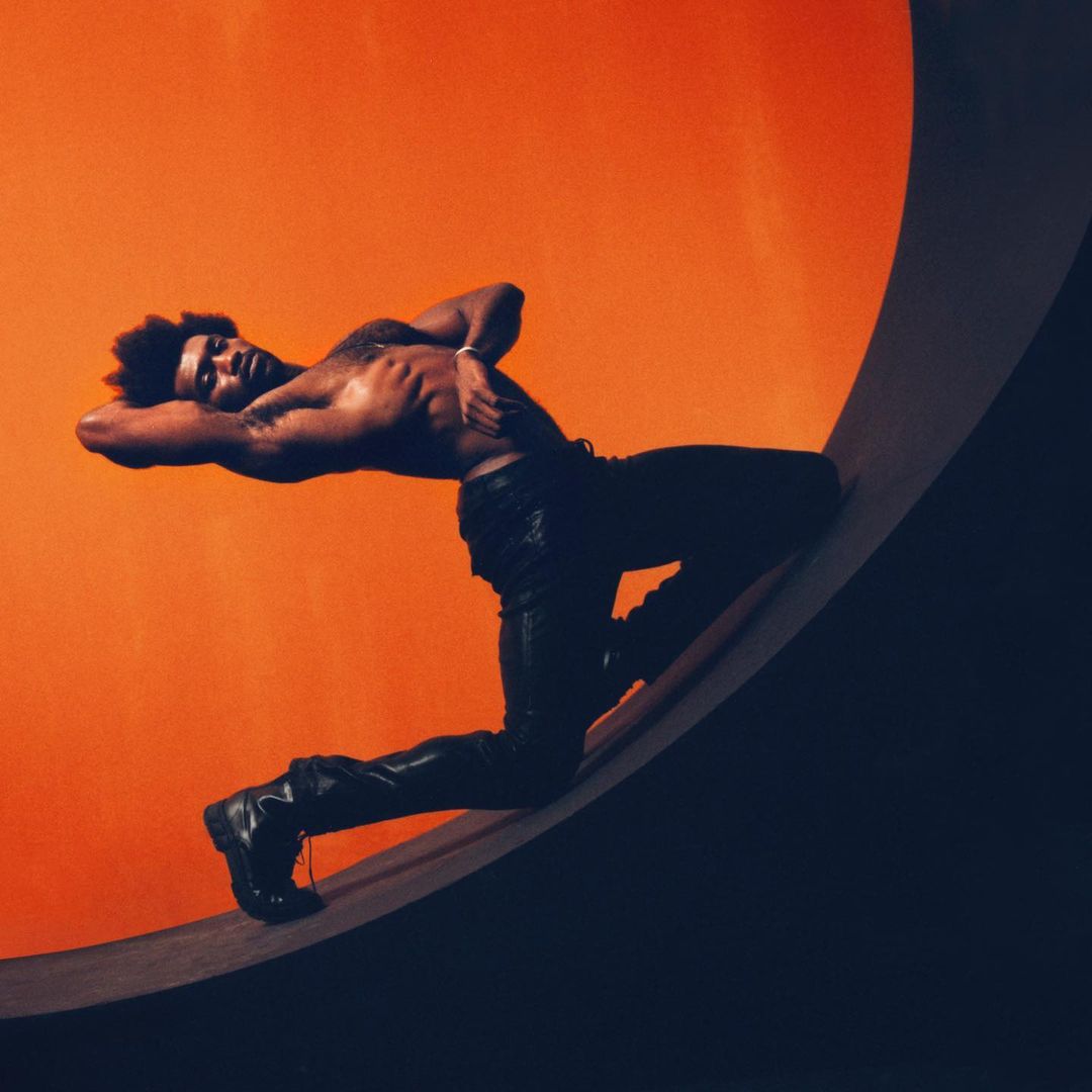 Artistic shot of a shirtless man in black pants posing on a gray and orange circular structure against an orange background in a studio setting.