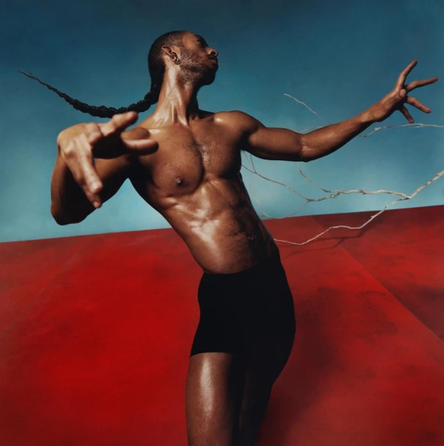 Athletic Black man with braided hair poses dynamically against a vibrant backdrop of blue sky and red geometric shapes in a studio setting.
