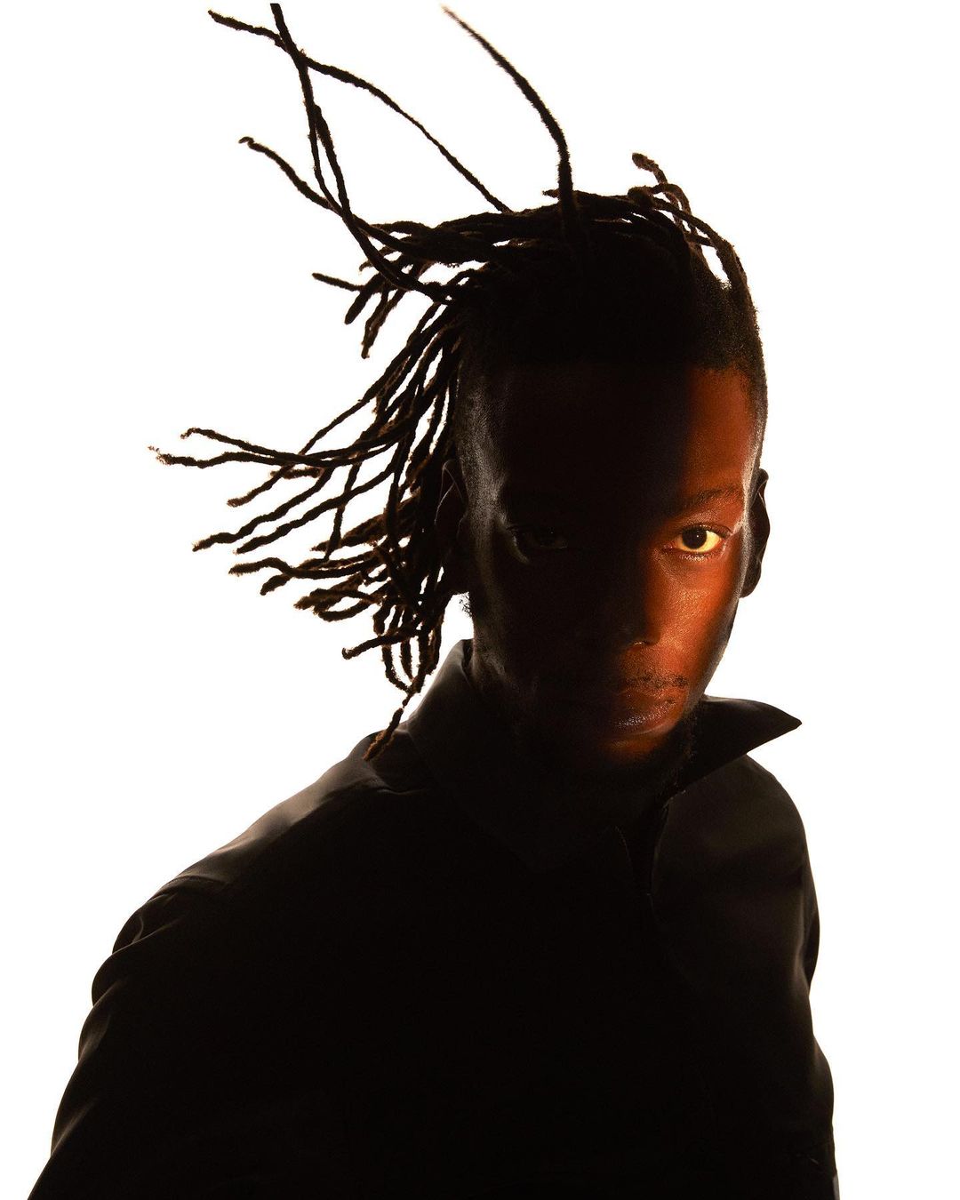 Dramatic portrait of a Black adult male with dreadlocks, glancing upward with a serious expression, half-shadowed against a bright white backdrop.