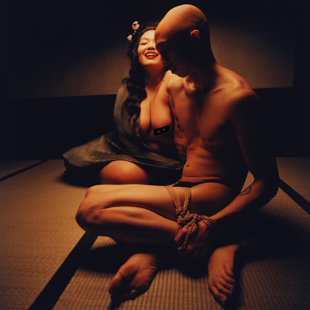 Striking image of an unclothed bald man and a woman in traditional clothing sitting on a tatami mat with dramatic lighting.