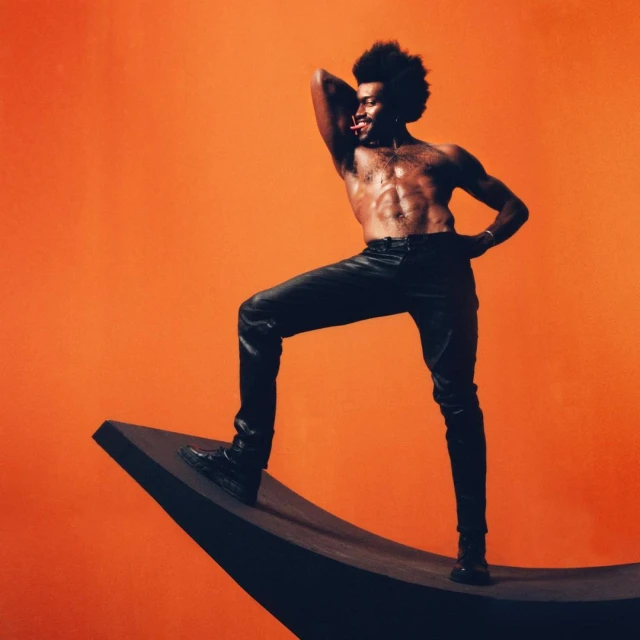 Shirtless black male model with afro, posing in black leather pants, balancing on modern black sculpture against an orange backdrop.