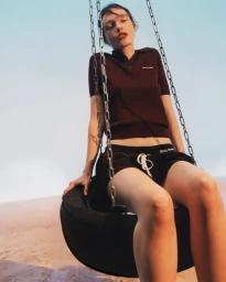 A woman in a brown polo shirt and black shorts seated on a suspended tire swing against a clear sky.