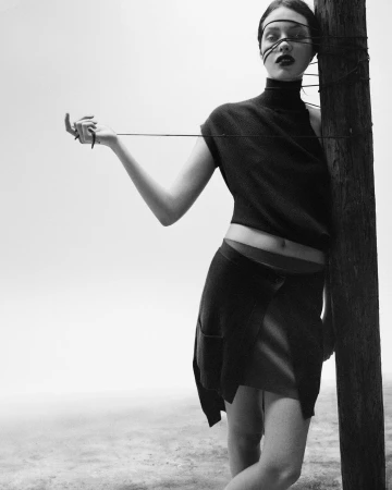 Model in dark sleeveless turtleneck and skirt wrapped in thin string tied to a wooden post against a pale background