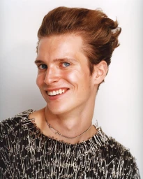 Portrait of a smiling white man with red hair wearing a black garment embellished with safety pins against a white background.