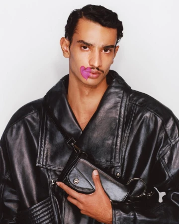 Man with a purple heart painted on his cheek, wearing a black leather jacket and holding a black leather shoulder bag.