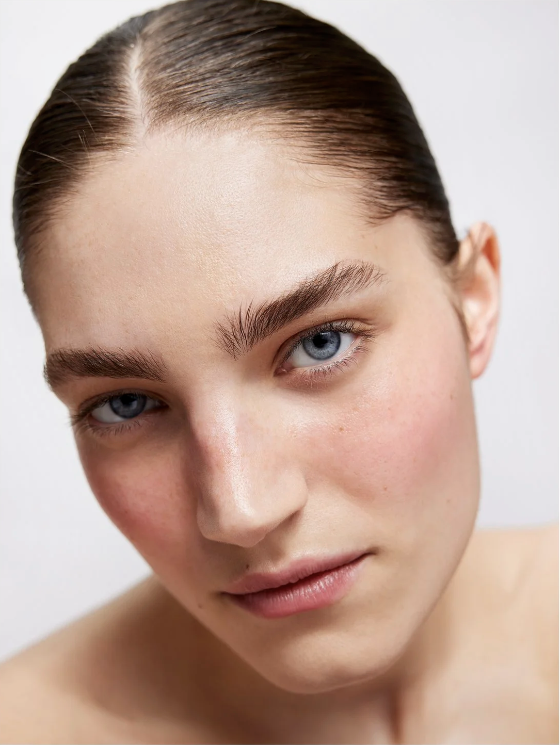 Close-up portrait of a young adult woman with fair skin, blue eyes, and slicked-back hair against a plain white background, beauty.