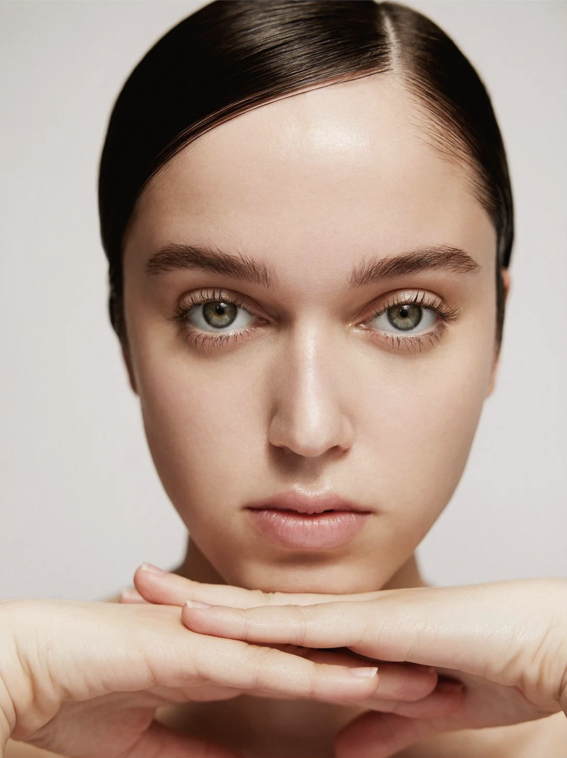 Close-up portrait of a young fair-skinned woman with slicked-back dark hair, and green eyes, posing with hands under her chin in a beauty shot.
