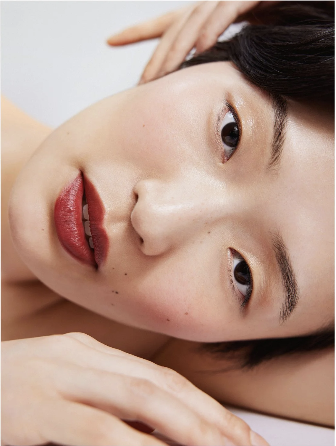 Close-up of an Asian woman with dark hair and red lipstick reclining with her hands near her face against a white background.