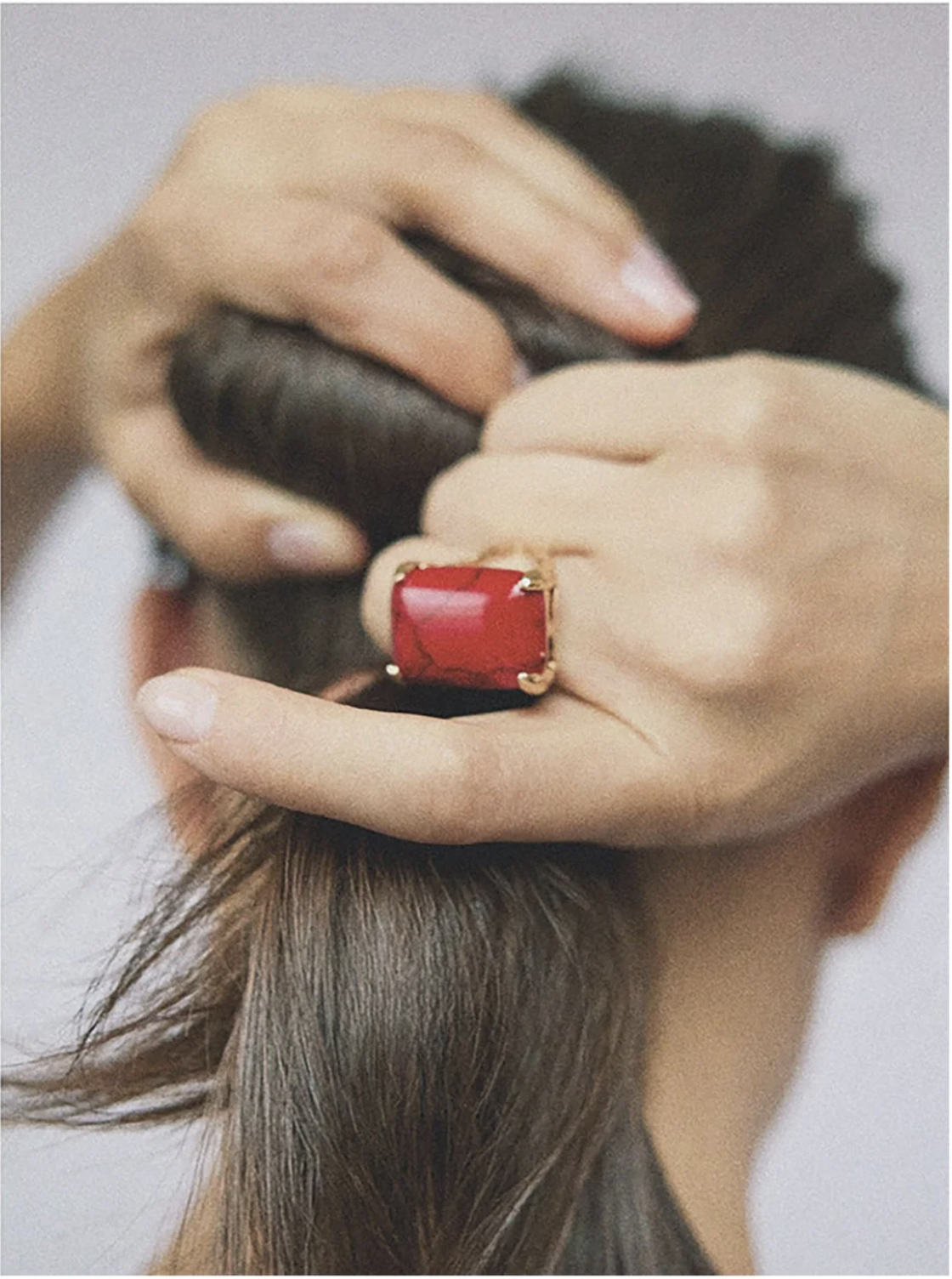 Close-up of a woman's hands securing her hair with a hair tie, showcasing a large red gemstone ring with a gold setting.