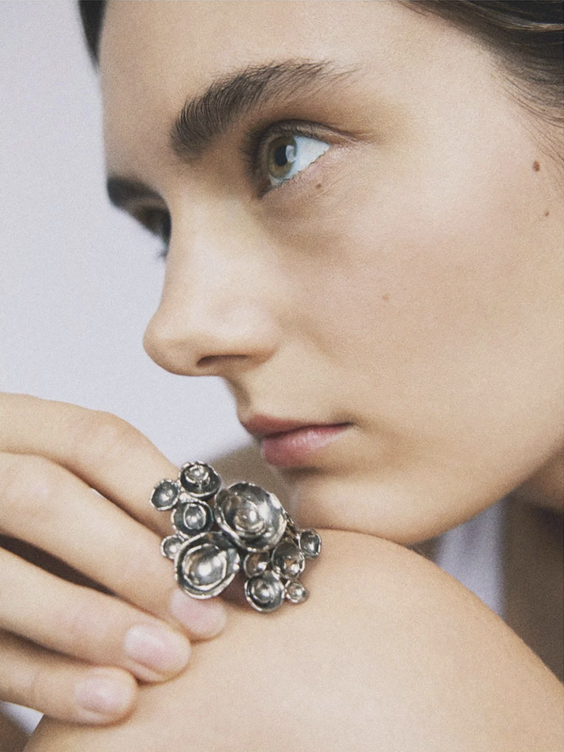 Close-up of a fair-skinned woman wearing a large, silvery, floral-shaped ring, posing against a neutral background, showcasing the beauty accessory.
