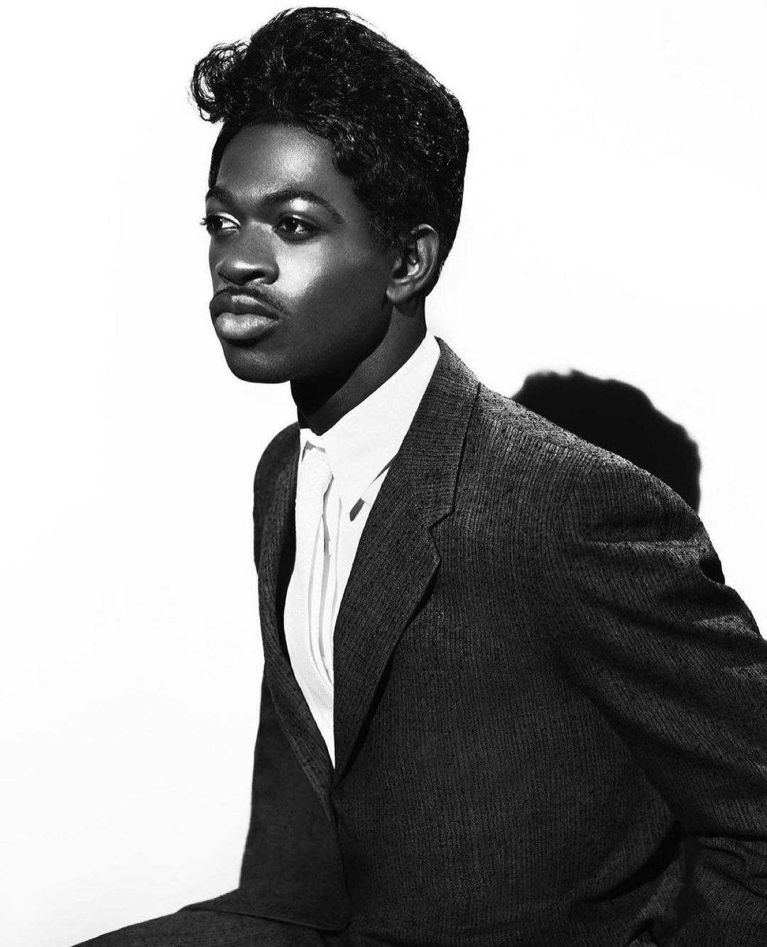 Black and white studio portrait of a man in a suit looking off to the side, with a bright background highlighting his features.