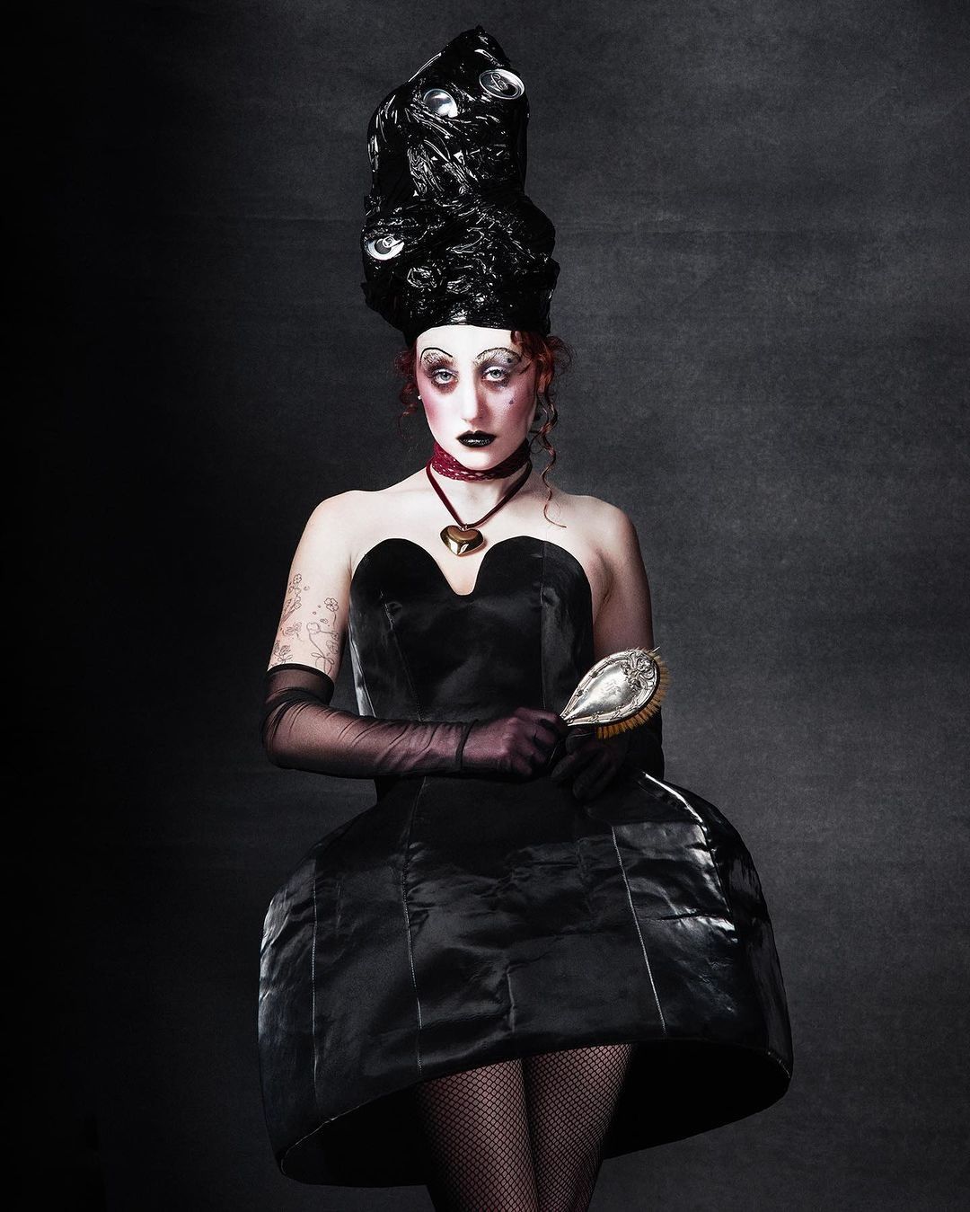 Woman in a dramatic black gown and theatrical makeup poses in a studio with a vintage hairbrush, creating a striking fashion editorial.