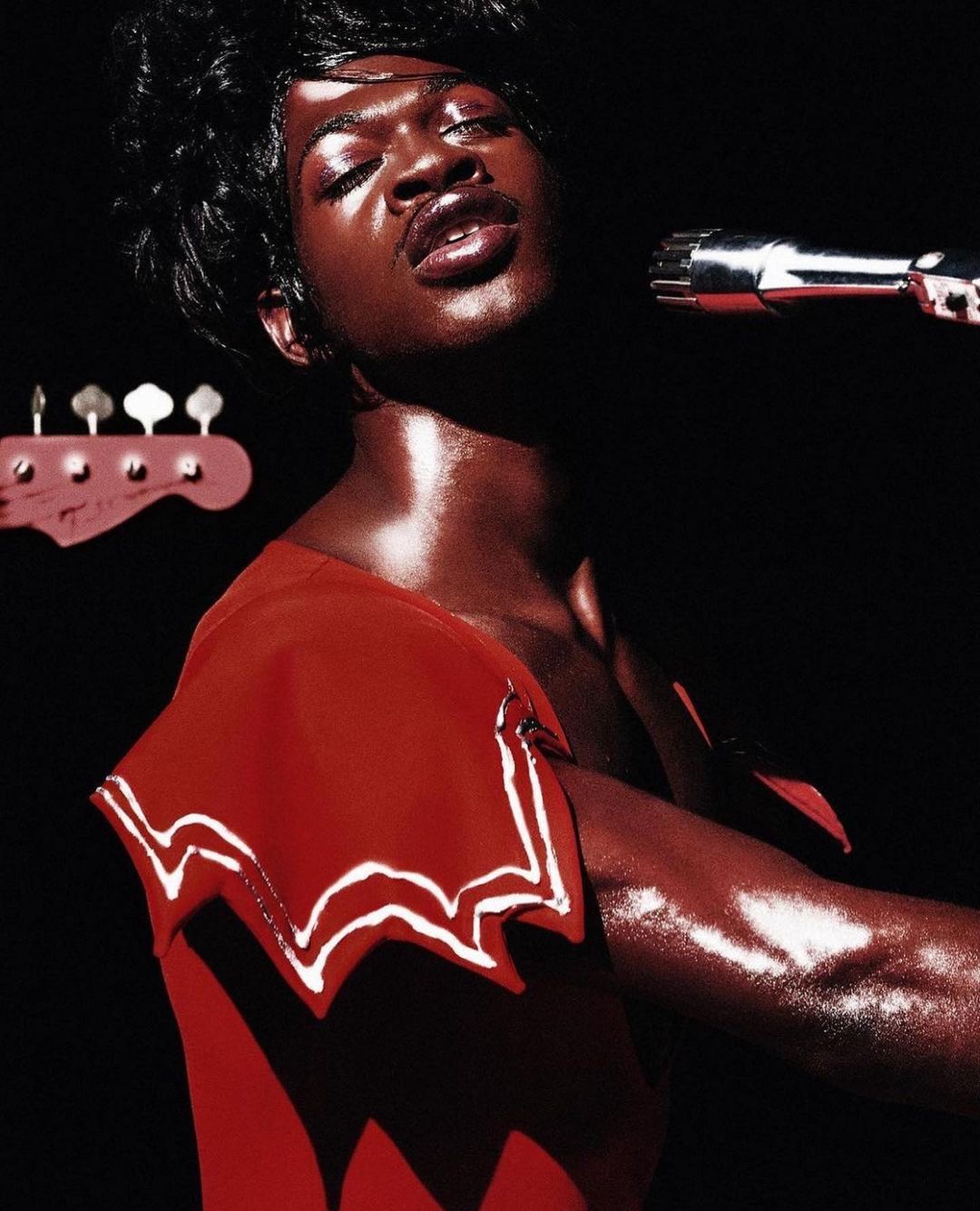 Lil Nas X sings into a microphone while wearing a red outfit, a bass guitar is visible in the background against a dark backdrop.