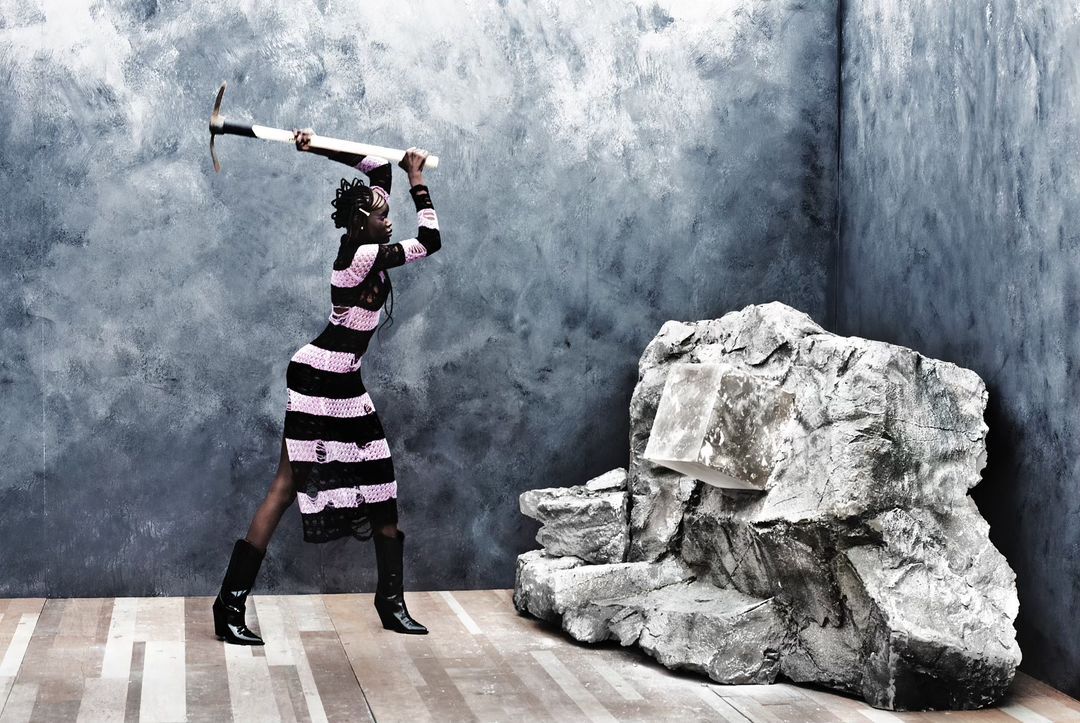 A woman in a black and pink dress wielding a pickaxe next to a rock formation, indoors against a textured gray and white wall backdrop.