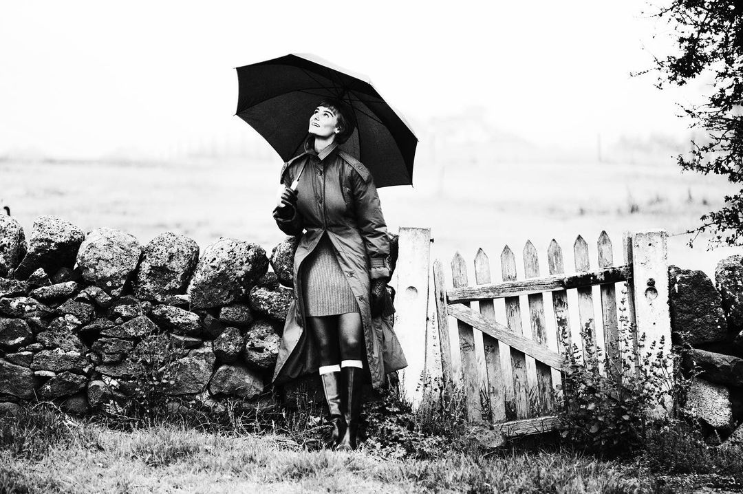 Black and white outdoor shot of a woman in a trench coat holding an umbrella, standing near a stone wall and wooden gate in a field.