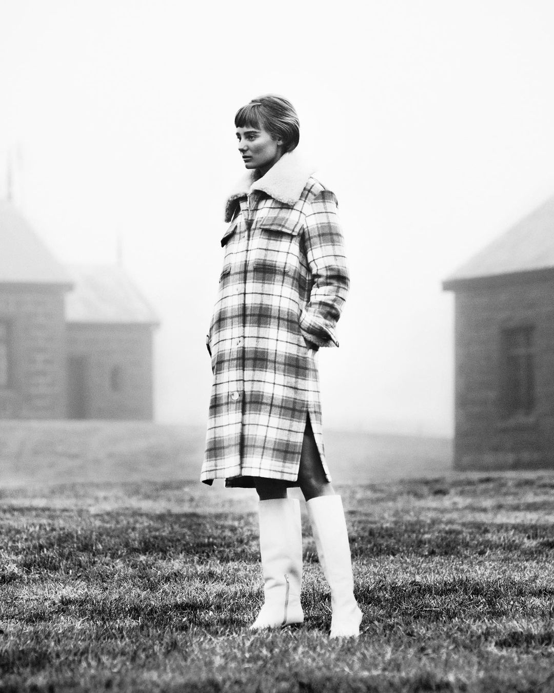 A stylish woman in a plaid coat and white boots stands in a field with blurred buildings in the background, captured in black and white.