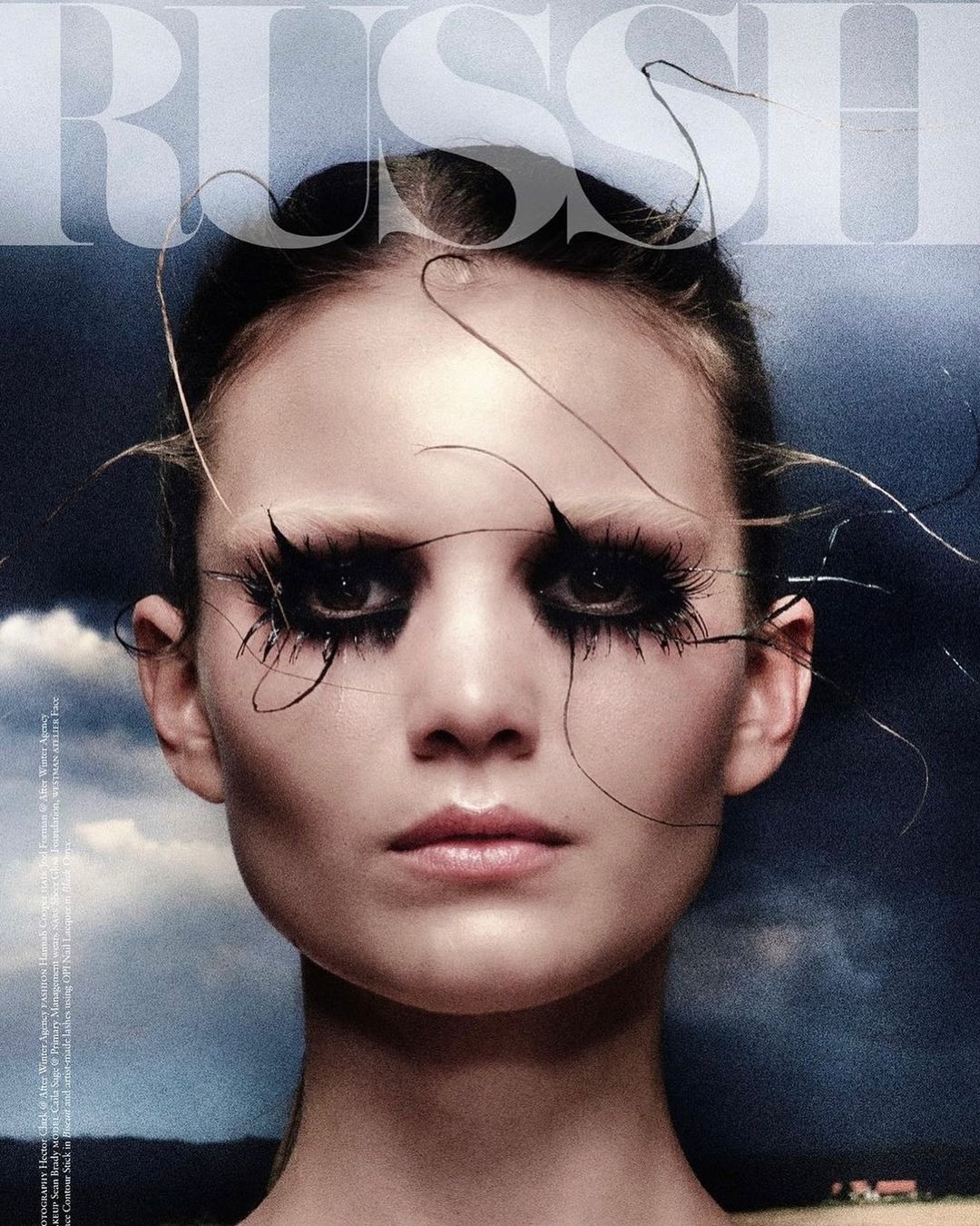 Fashion portrait of a woman with dramatic makeup and long eyelashes, shot against a moody sky with hints of landscape in the distance.