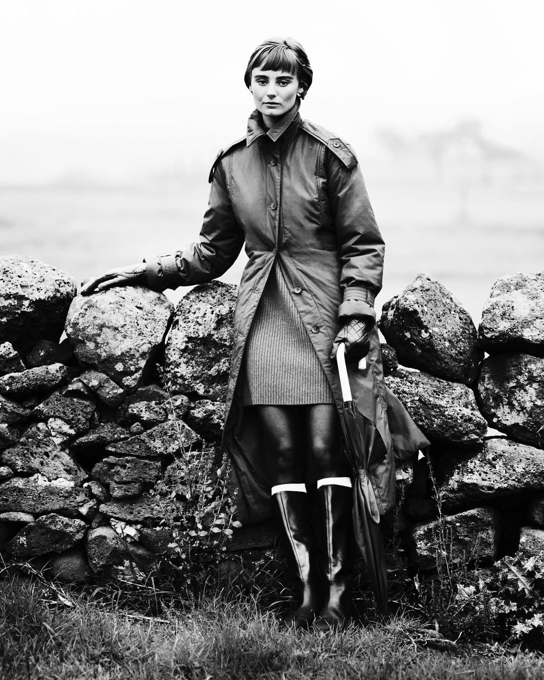 Woman in trench coat, dress, and rain boots poses near stone wall in black and white, holding an umbrella in a rural outdoor setting.