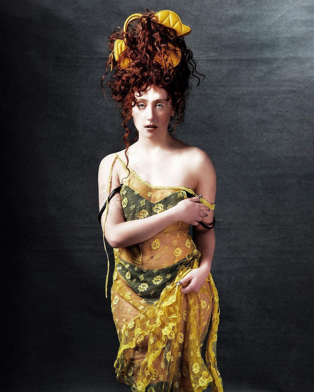 Woman poses in a sheer yellow floral dress with curly auburn hair and a yellow headpiece against a mottled dark background in a portrait.