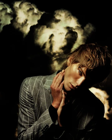 Portrait of a blond male fashion model wearing a pinstripe suit jacket, posing with hands near face in a dramatic light, against a dark backdrop.