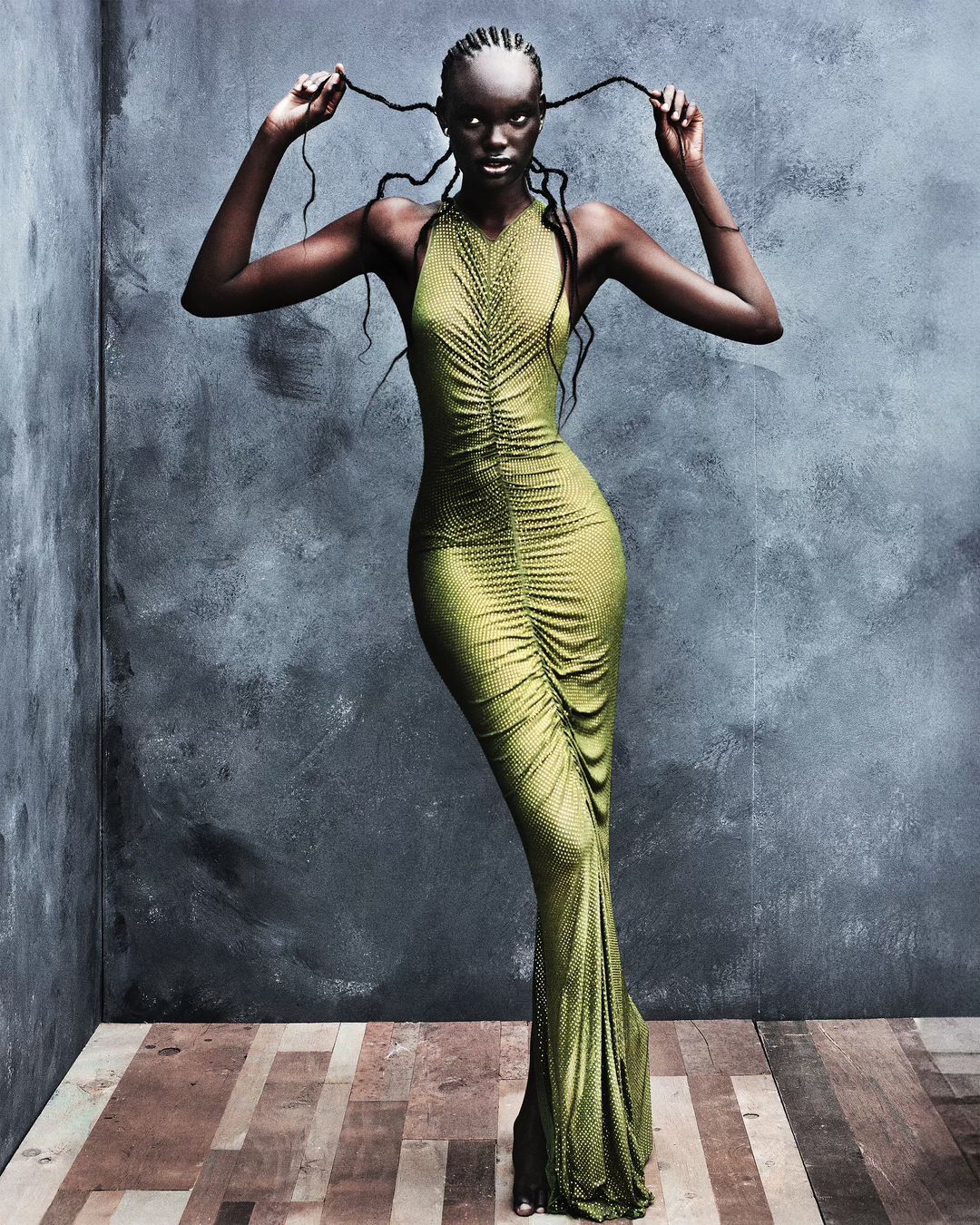 Elegant woman in a floor-length olive green gown poses against a textured grey wall, her braids playfully framing her face in a fashion shot.