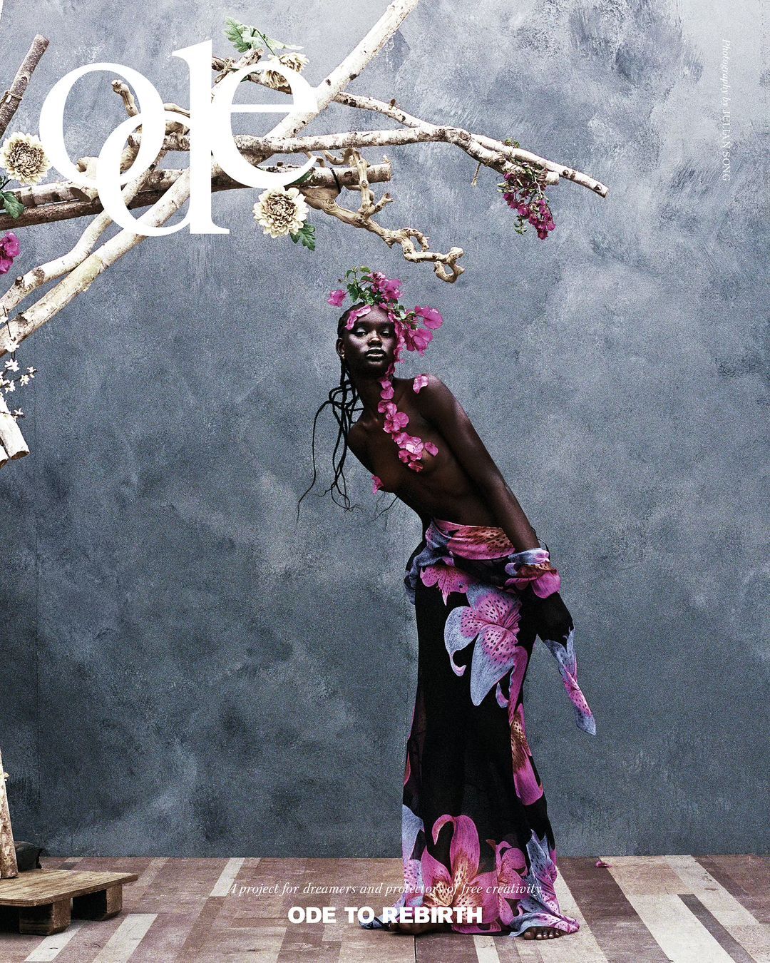 Fashion editorial shot of a black female model adorned with pink flowers, wearing a floral-patterned dress, posing against a grey wall.