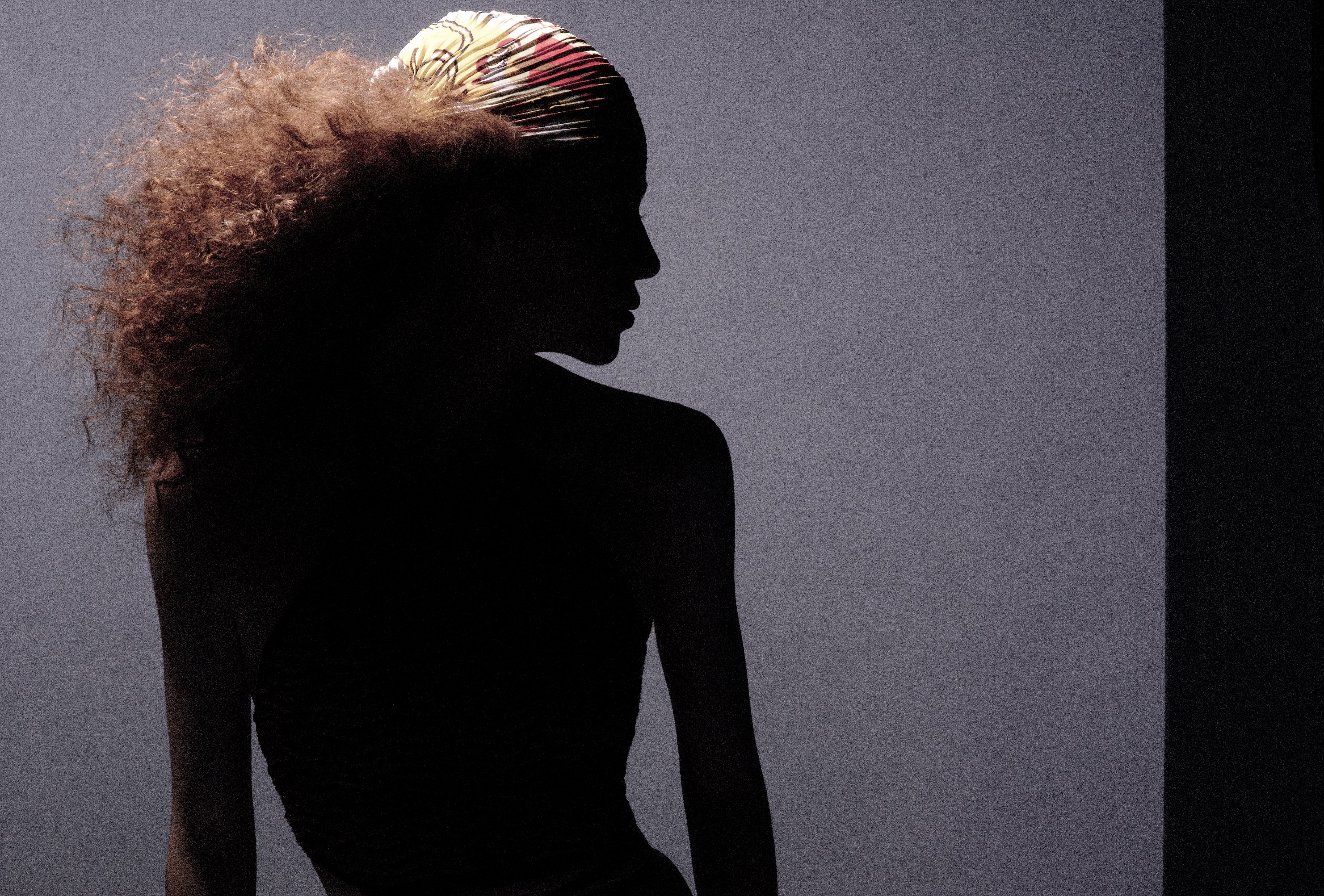 Silhouette of a woman in shadow wearing a patterned headscarf, gazing toward the light. Neutral gray background.