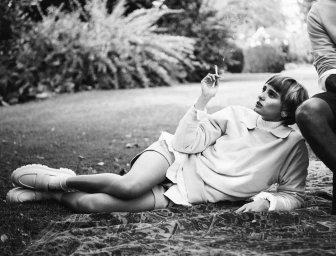 Woman in light-coloured sweatshirt and shorts reclining on a patterned blanket outdoors holding a cigarette