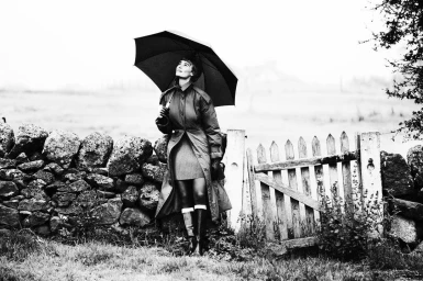 Woman holding a black umbrella standing by a stone wall and wooden gate outdoors wearing a belted trench coat and knee-high boots.