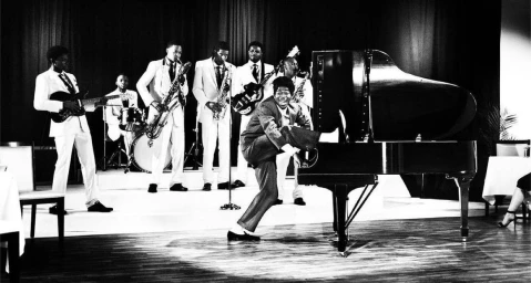 Black and white portrait of a performer kicking a grand piano while a band in white suits plays in the background