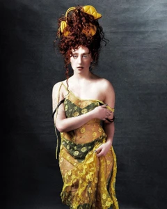 Portrait of Chappell Roan with elaborate voluminous red hair and yellow bag accessories, wearing a sheer patterned yellow lace dress.