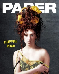 Chappell Roan with voluminous red hair and yellow handbags pinned in it wearing a patterned yellow top against a dark textured background