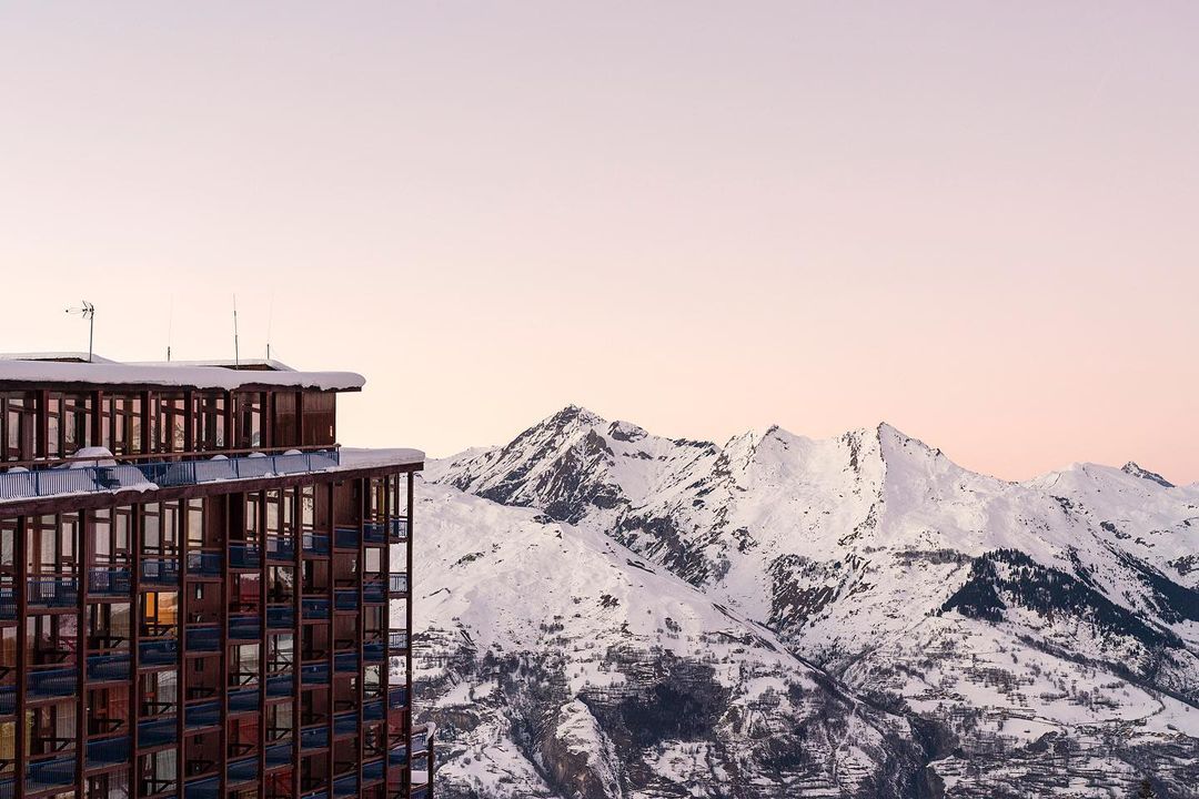 High-rise building stands next to snow-covered mountain range under a pastel sky, creating a scenic alpine view with architectural contrast.