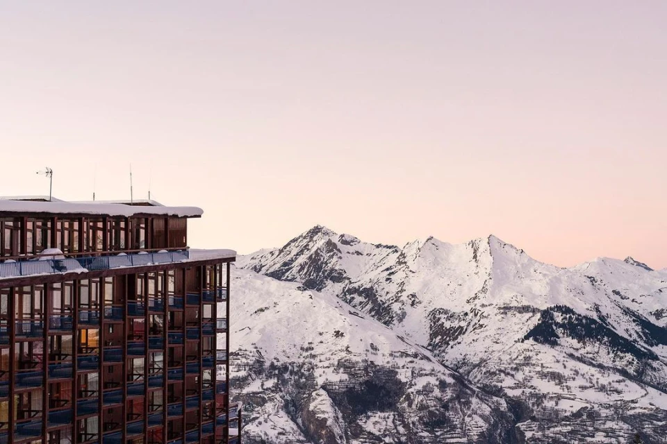 High-rise building stands next to snow-covered mountain range under a pastel sky, creating a scenic alpine view with architectural contrast.