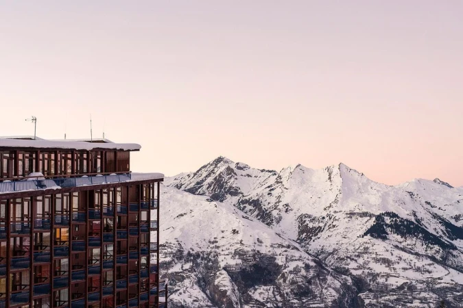 High-rise building stands next to snow-covered mountain range under a pastel sky, creating a scenic alpine view with architectural contrast.
