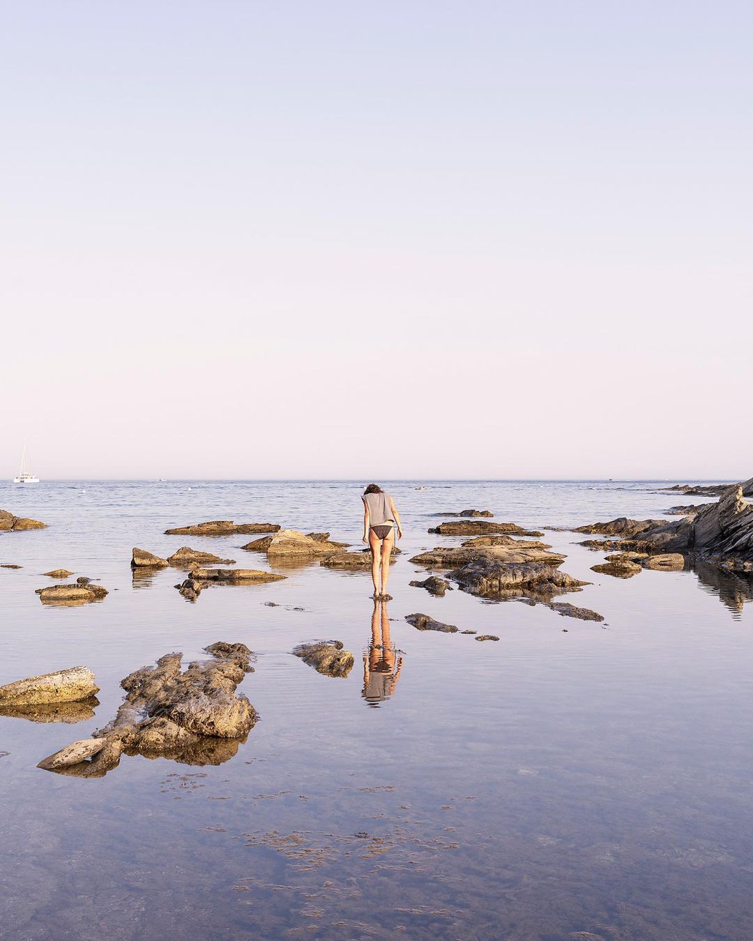Woman in swimwear reflected in shallow water with rocks on the shore. Soft light on the ocean, sky, and distant boat.