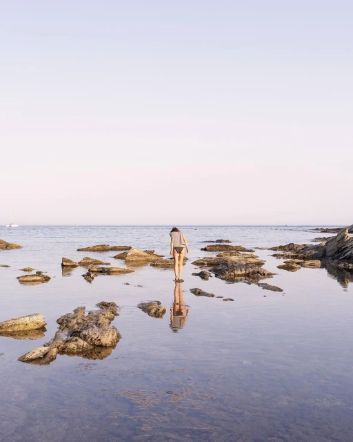 Woman in swimwear reflected in shallow water with rocks on the shore. Soft light on the ocean, sky, and distant boat.