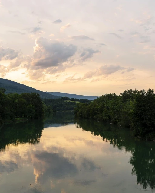 Scenic landscape featuring a calm river reflecting the sky, framed by green trees and distant mountains at dusk or dawn.