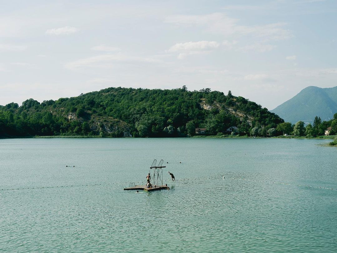 Scenic view of a lake with a floating platform and people diving, surrounded by lush green hills and a clear sky. Outdoor recreation.