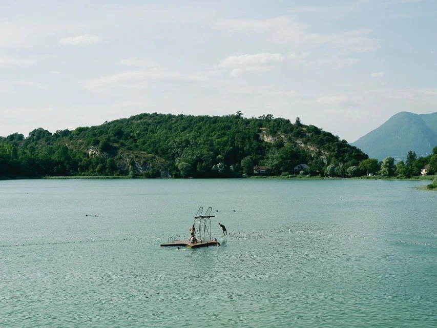 Scenic view of a lake with a floating platform and people diving, surrounded by lush green hills and a clear sky. Outdoor recreation.