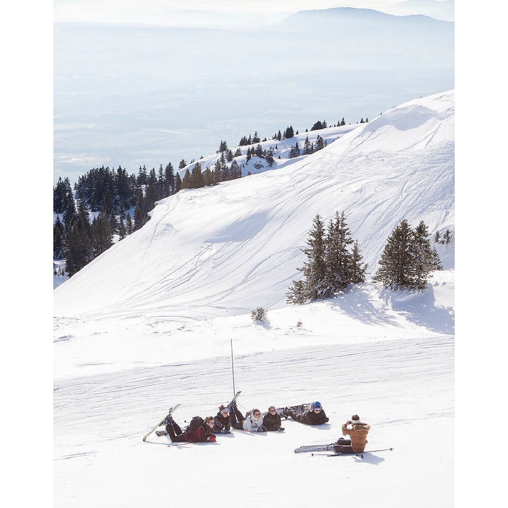 A group of skiers relaxes on a snowy mountain slope. Some are reclining, while one individual sits and takes a photo against a mountain backdrop.