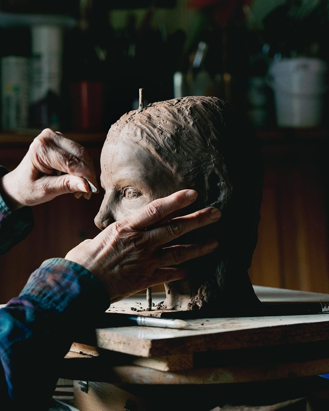 Adult makes clay sculpture bust indoors, showing creative process, hand-modeling detail, dark wood setting, and art studio background.