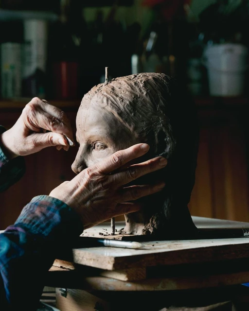 Adult makes clay sculpture bust indoors, showing creative process, hand-modeling detail, dark wood setting, and art studio background.