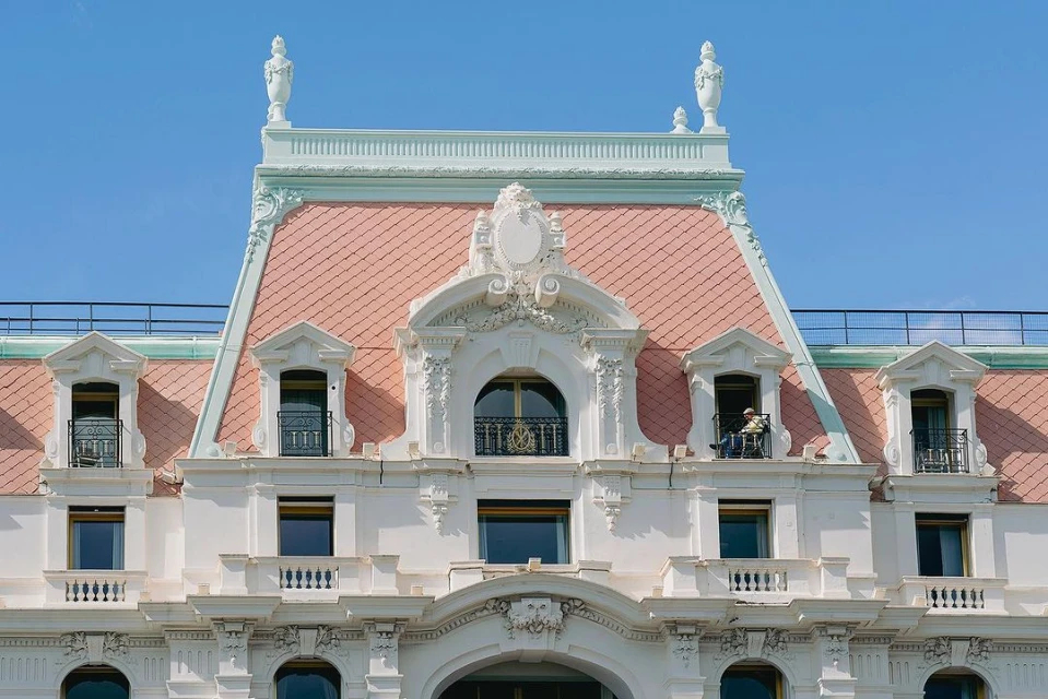 Architectural detail of a white building facade with a pink tiled roof and several windows on a bright day under a clear blue sky.