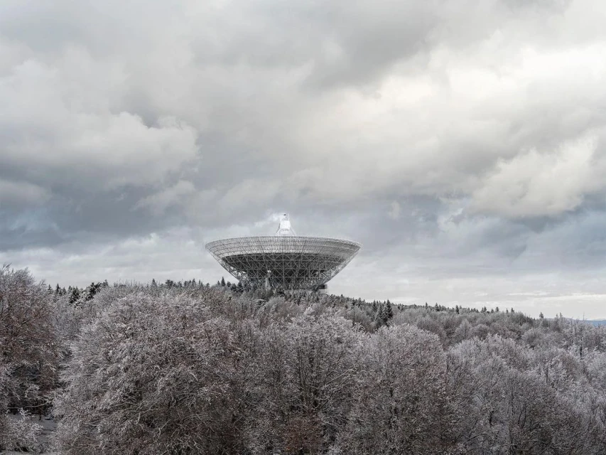 Wide shot of large radio telescope on a hill surrounded by snowy trees under a cloudy sky, reflecting cold, quiet technology and nature.